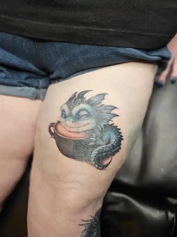 Color tattoo on thigh of a sleepy dragon character curled inside a coffee mug