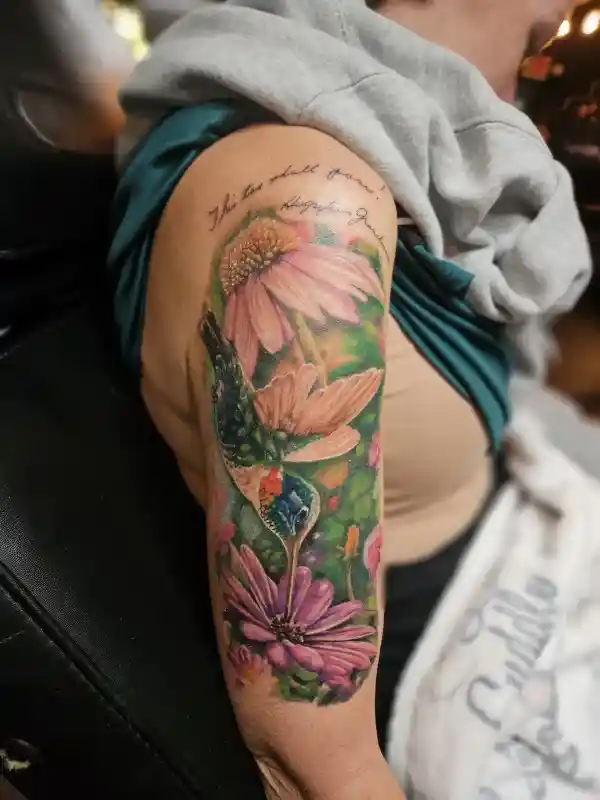 Color tattoo on upper arm of a hummingbird among pink coneflowers with script lettering