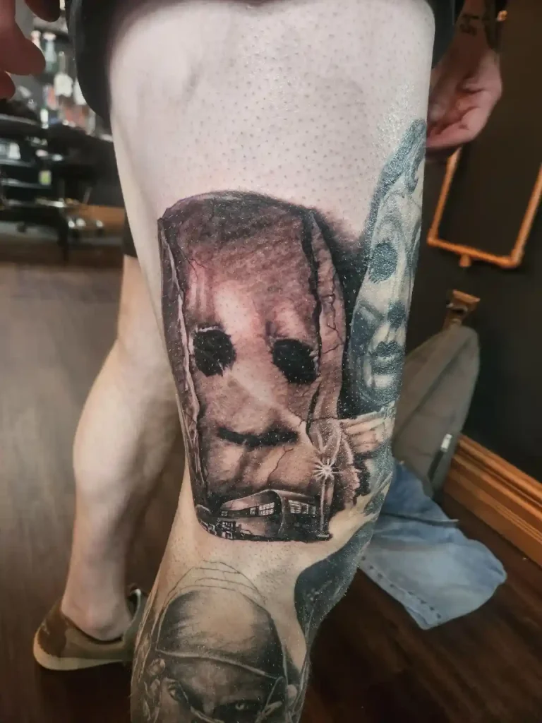 Black and grey tattoo on leg of a masked horror character with a house in the background
