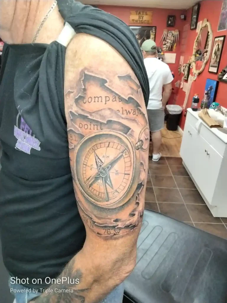 Black and grey tattoo on upper arm of a compass and map with coordinates and lettering