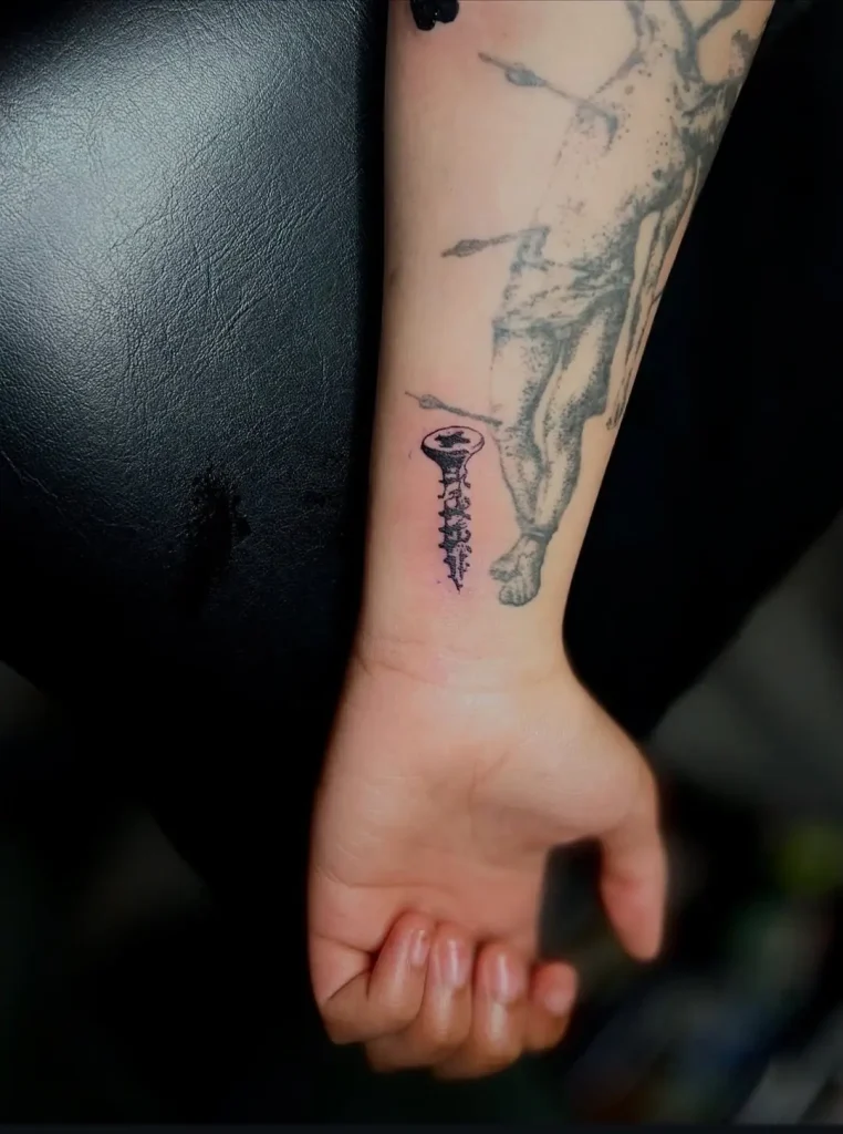 Small black ink screw tattoo on wrist, fine line detail by Tainted Flesh Body Art North artist