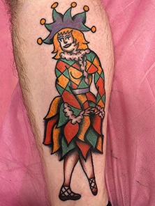 Traditional style jester tattoo on calf featuring a diamond-patterned harlequin costume in green, orange and red with a purple jester hat