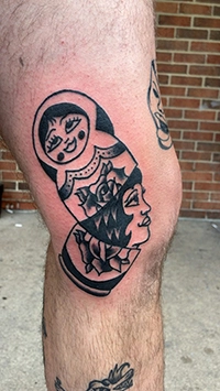 Black linework nesting doll and face tattoo on the knee