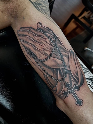 Black and grey praying hands tattoo with a rosary and cross on the forearm