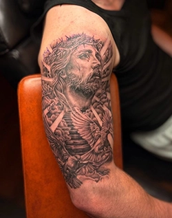 Black and grey religious tattoo of a robed figure with a crown of thorns on the upper arm