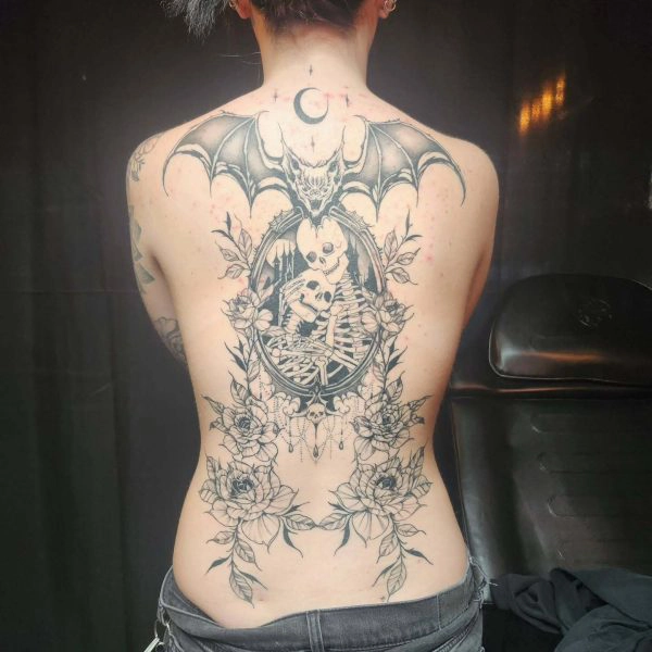 Black and grey back tattoo featuring a bat, skeleton couple in an oval frame, and roses