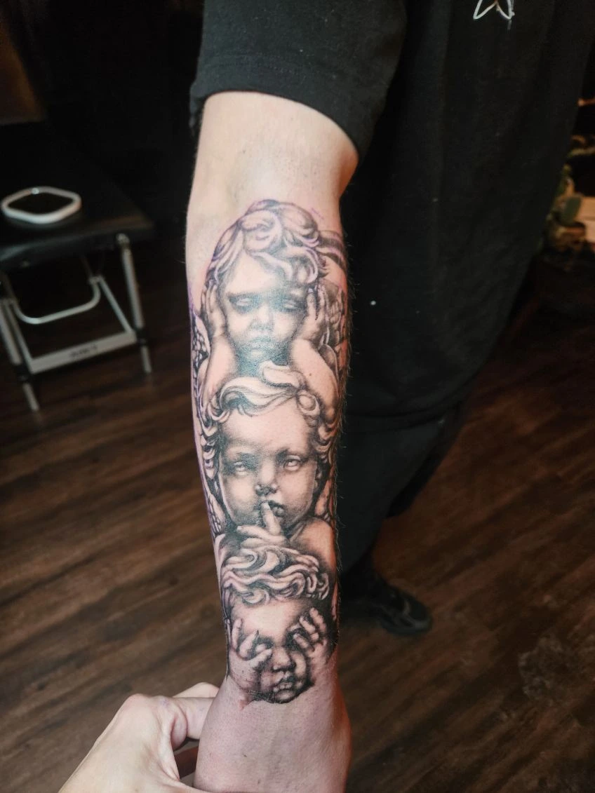 Black and grey forearm tattoo of three cherub angels in "hear no evil, speak no evil, see no evil" pose with realistic statue-like shading