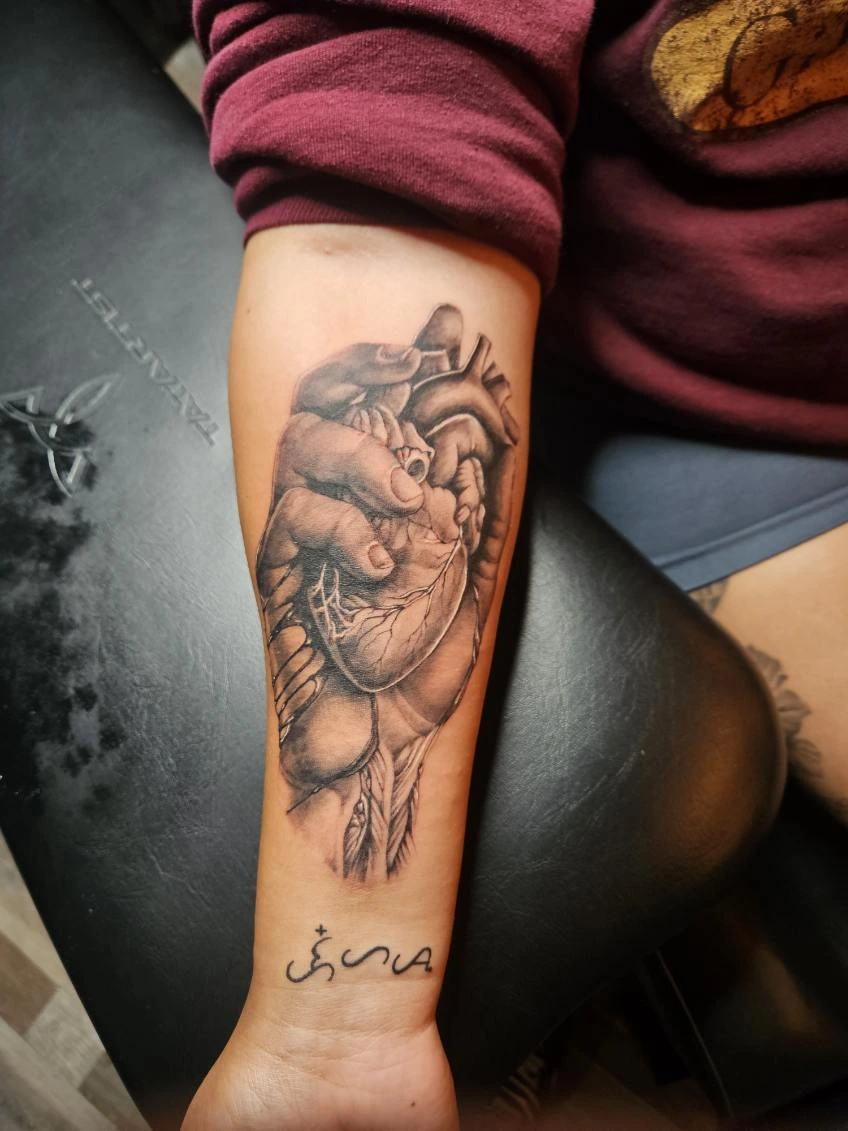 Black and grey forearm tattoo of an anatomical heart with clenched fist detail, fine vein linework and a small feather accent below
